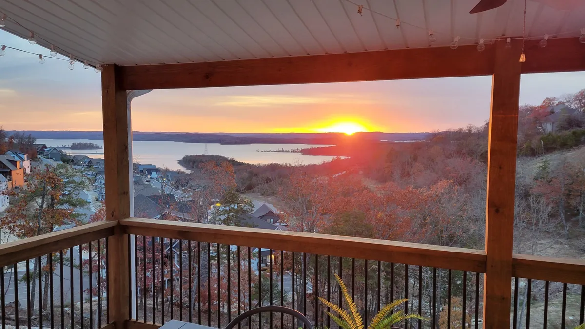 A serene lake view from the deck, with a sunset casting warm light over the hills, wooden railing, and a table adorned with decorative plants. The setting is perfect for alfresco dining or simply enjoying the scenery.