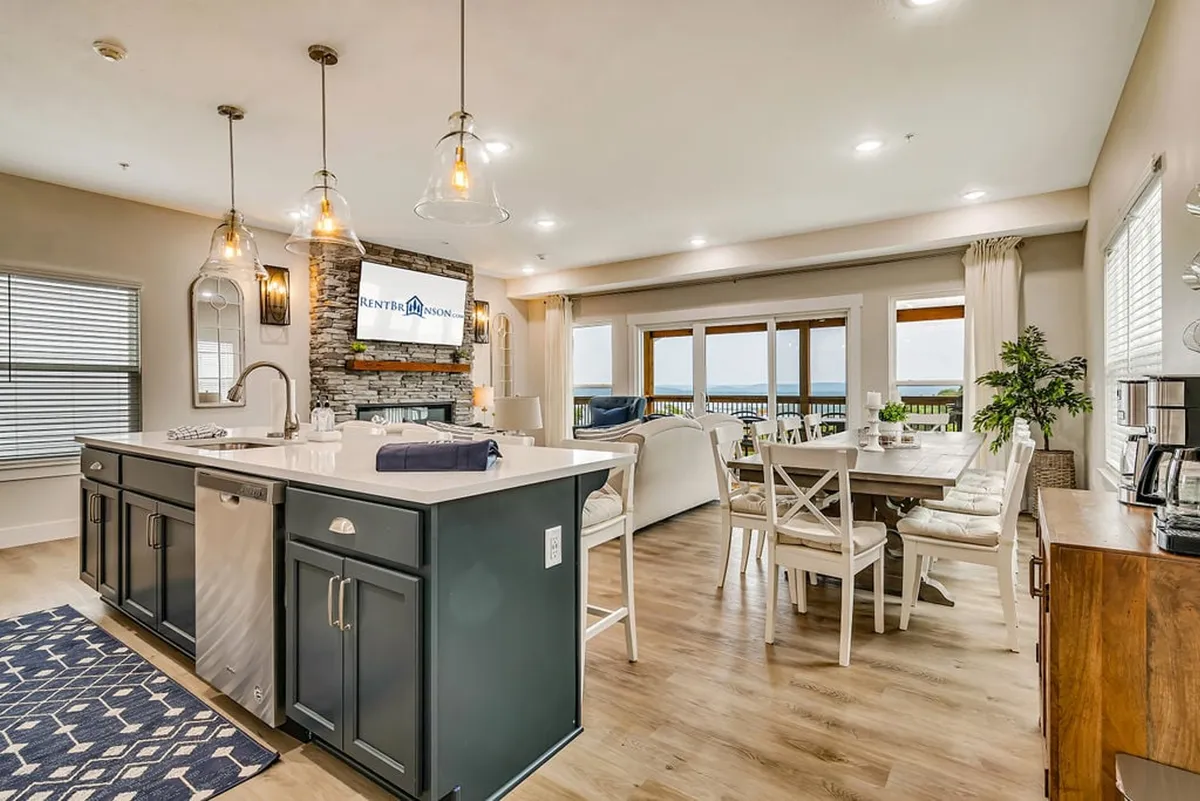 The hero image showcases an inviting open-concept living space featuring a spacious kitchen island, a dining table set for gatherings, large windows flooding the room with natural light, and a beautiful stone fireplace. The scene suggests comfort, warmth, and a perfect setting for family meals, cozy evenings, and enjoying panoramic views of Table Rock Lake.