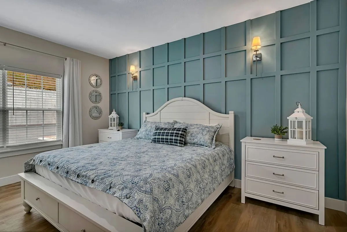 A serene bedroom featuring a plush king-sized bed adorned with patterned bedding, flanked by nightstands and lamps. The blue accent wall, decorative mirrors, and soft lighting create a cozy and inviting atmosphere, perfect for unwinding after a day by the lake.