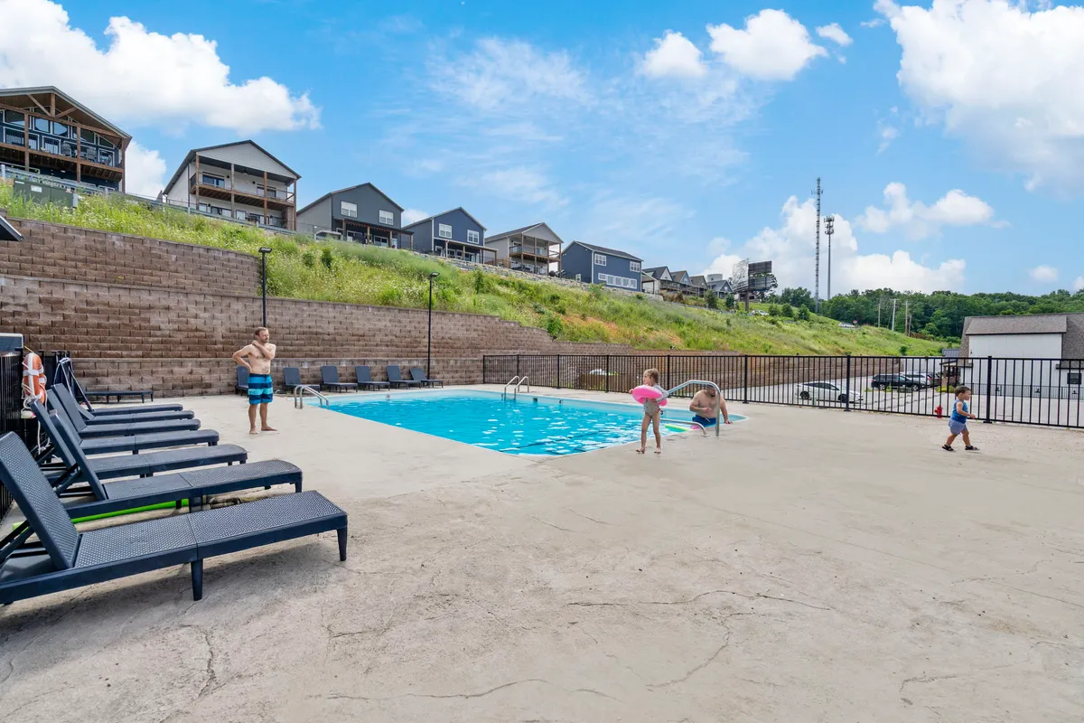An inviting outdoor swimming pool area with children laughing and playing in the water while adults relax on poolside lounge chairs. The scene is bright and energetic, with the pool framed by sun loungers and a spacious deck. The overall ambiance suggests cheerful family gatherings and summer fun.
