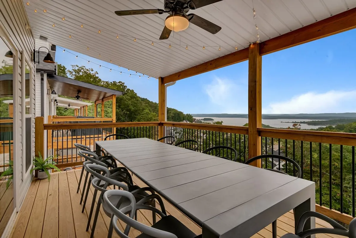 A long outdoor dining table on a deck, set beneath string lights and overlooking a beautiful lake. The space is designed for gatherings and leisurely meals, with the shimmering water and mountains visible in the background, enhancing the overall sense of tranquility and luxury.