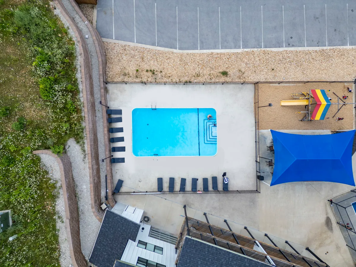 A vibrant exterior space featuring a swimming pool, sun loungers, and a colorful playground area under a shade structure. The scene captures families spending time together—children playing in the playground while adults unwind by the pool. The lush landscaping and sunny weather make the environment feel welcoming and fun for all ages.