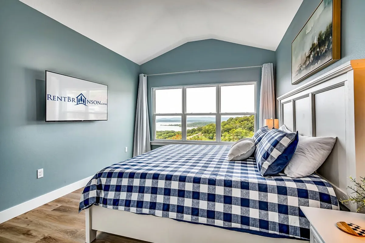 A comfortable bedroom with a checkered duvet on a king-sized bed, large windows offering scenic views, a wall-mounted TV, and tasteful artwork above the bed. Natural light fills the room, enhancing the tranquil and restful mood.
