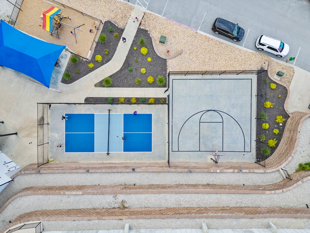 A bustling outdoor scene with tennis and basketball courts, a playground area, and landscaped gardens. People are seen playing sports, children are active on the playground, and residents are strolling along paths, reflecting an energetic and communal spirit.
