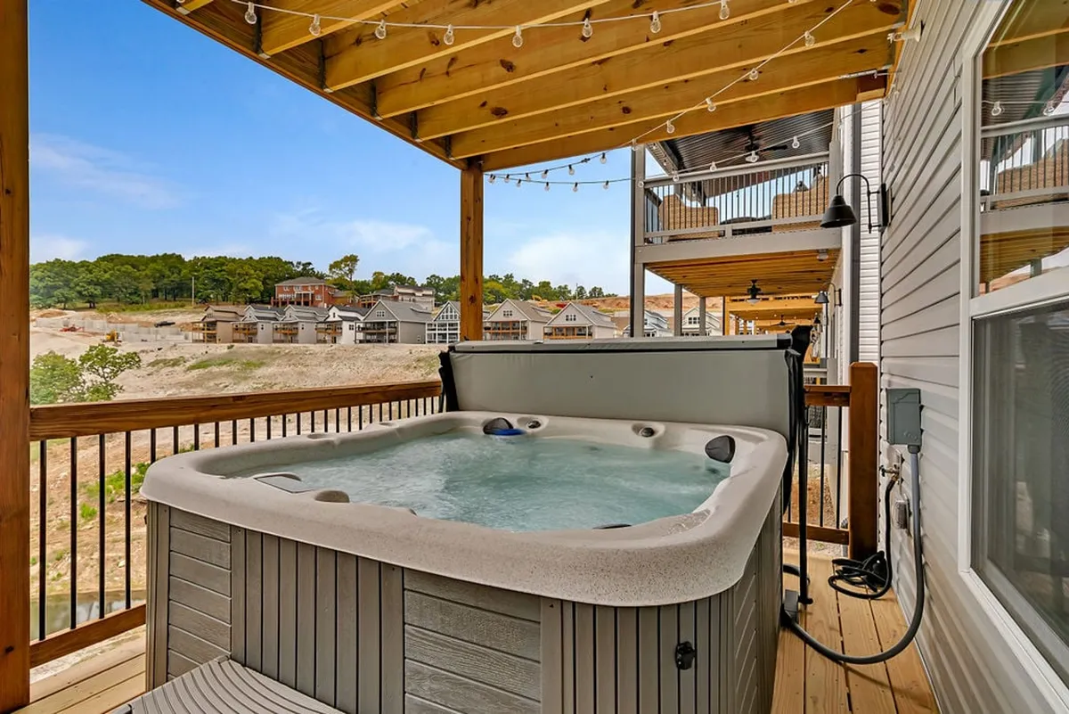 A private hot tub set on a wooden deck, surrounded by string lights that create a cozy and inviting evening atmosphere. The view from the deck showcases tranquil natural scenery, including glimpses of lake and mountain landscapes. The space is clearly designed for relaxation and socializing, inviting guests to unwind while soaking in the hot tub or enjoying the peaceful outdoor setting.