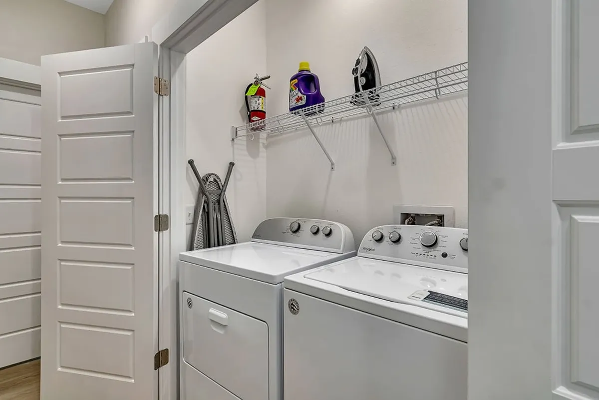 A tidy laundry area featuring a washing machine, drying machine, organized shelves with cleaning supplies, and an ironing board. The practical setup suggests convenience for longer stays and cost savings on laundry services.