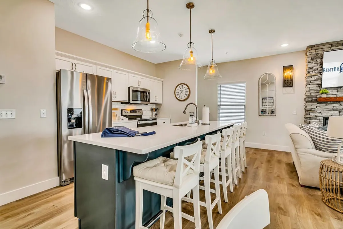 A bright, modern kitchen featuring an island counter with bar stools, pendant lights, stainless steel appliances, and a distinctive stone accent wall. The setup is perfect for cooking, entertaining, and casual dining.