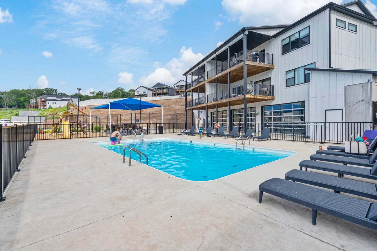 An inviting outdoor pool scene featuring sun loungers, a nearby playground, balconies of adjacent buildings, and people enjoying the pool area. The atmosphere is joyful, with activities like swimming, sunbathing, and socializing taking place.
